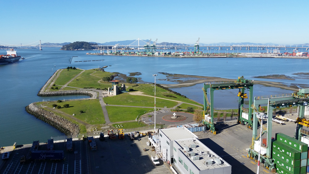 Aerial photo of the Port of Oakland at low tide. An industrial bulding on a concrete area is in the foreground with a green iron beam structure to the lower-right. A grassy penninsula extends into the water with criss-crossed roads. In the center right, water is over low land areas. In the upper portion is another structure resembling a very long barge or a man made penninsula with industrial constructions. A bridge and mountains are in the distance.